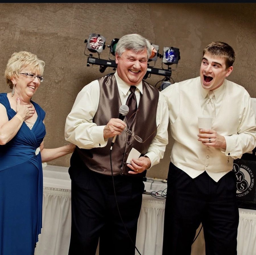 A man giving a toast with a microphone, laughing, beside his wife at a wedding reception.