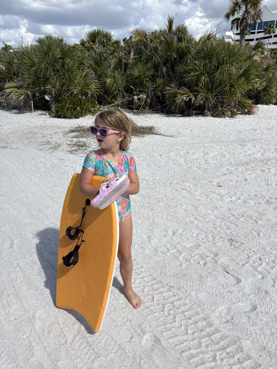 A young girl in sunglasses holding an orange bodyboard on white sand.