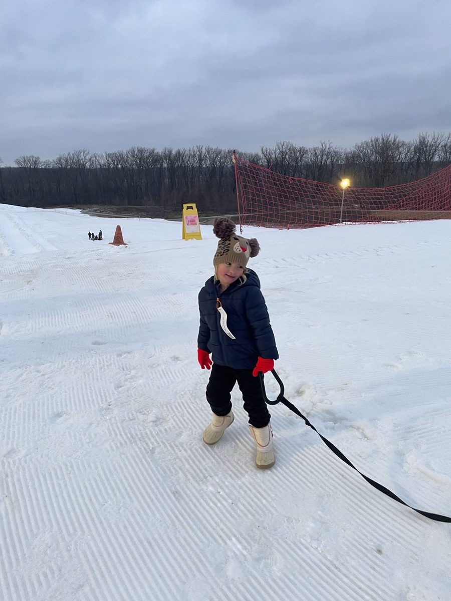 A young girl in a winter coat and hat pulling a snow tube across a snowy hill.