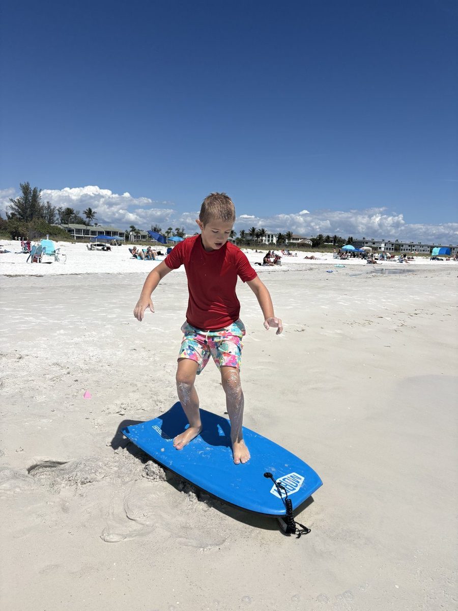 A boy in a red shirt standing on a blue bodyboard on a Longboat Key beach.