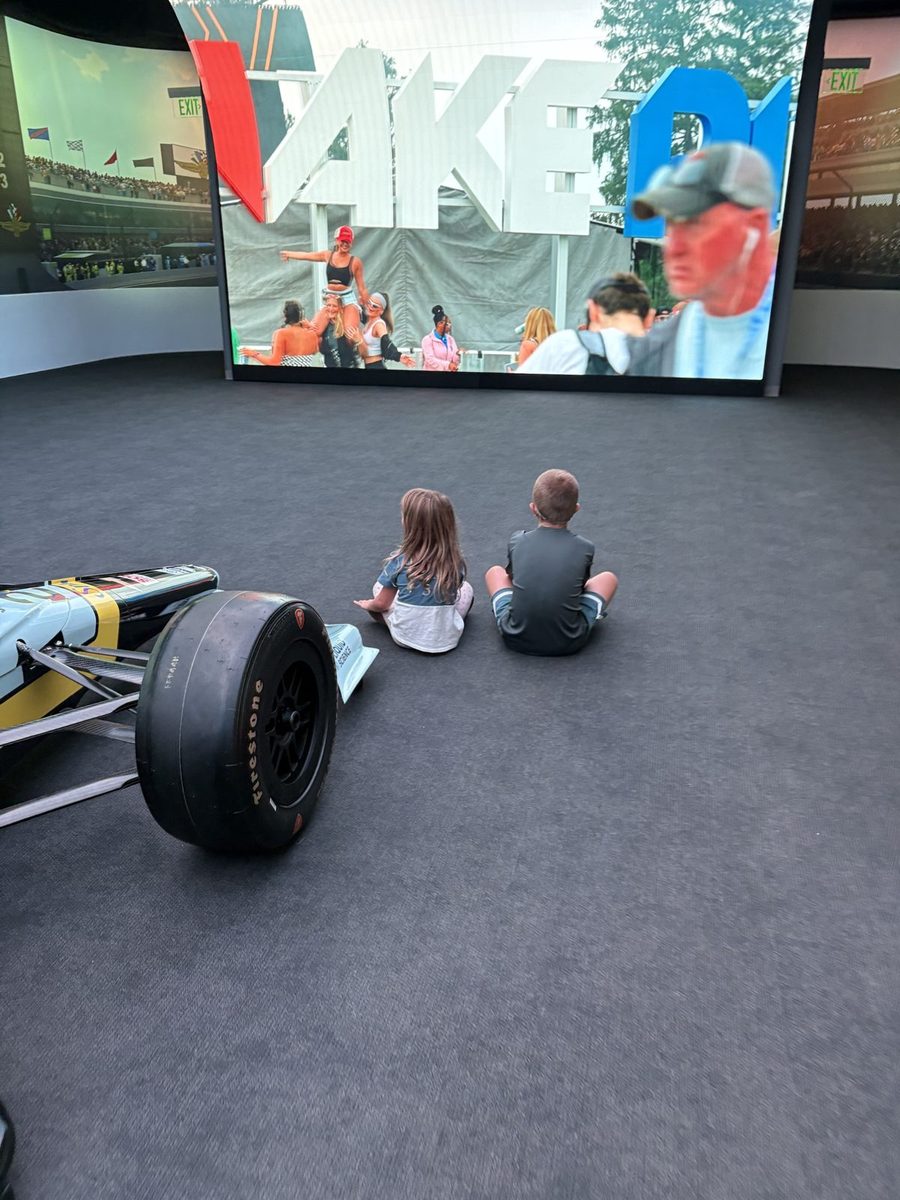 Two children sitting on the floor of a museum next to a race car, watching a large screen.
