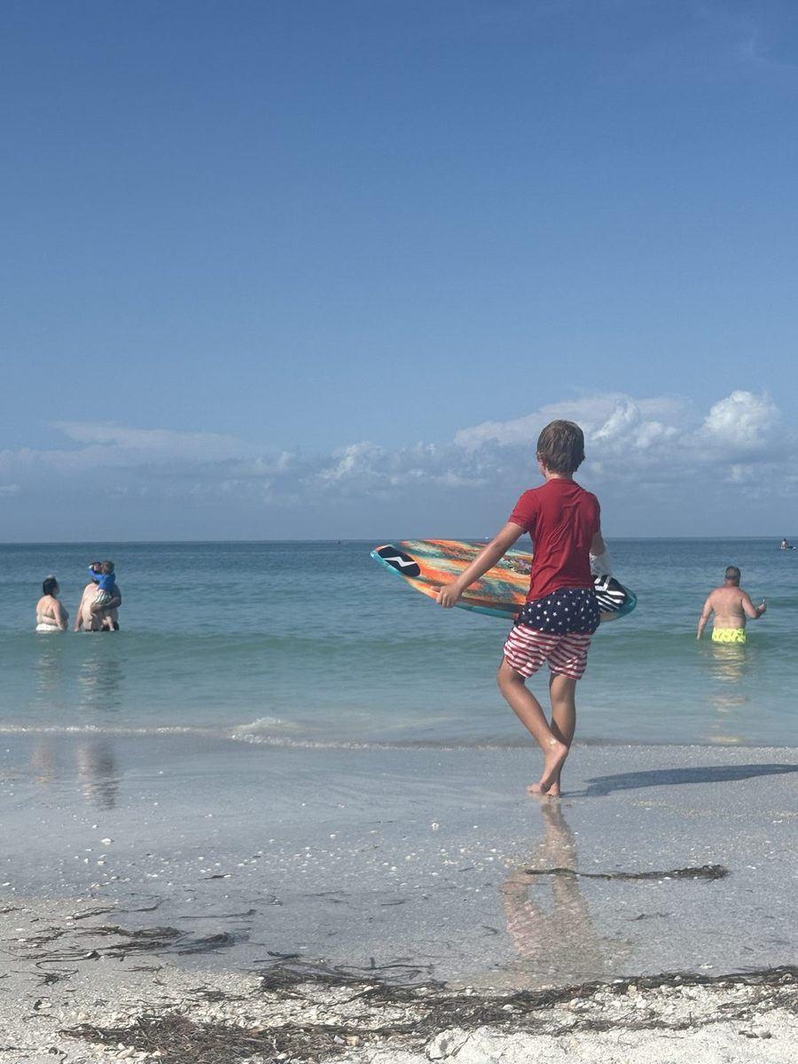 A boy in flag-print shorts carrying a skimboard into the Gulf.